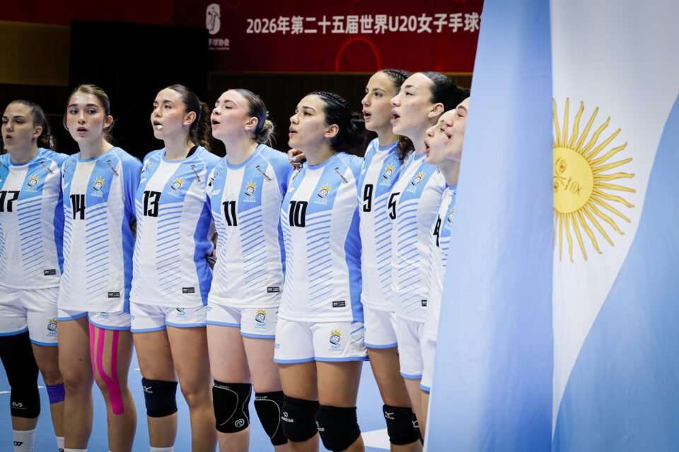 A team of athletes in light blue and white uniforms stands in a line, singing with their mouths open, beside a large flag of Argentina.